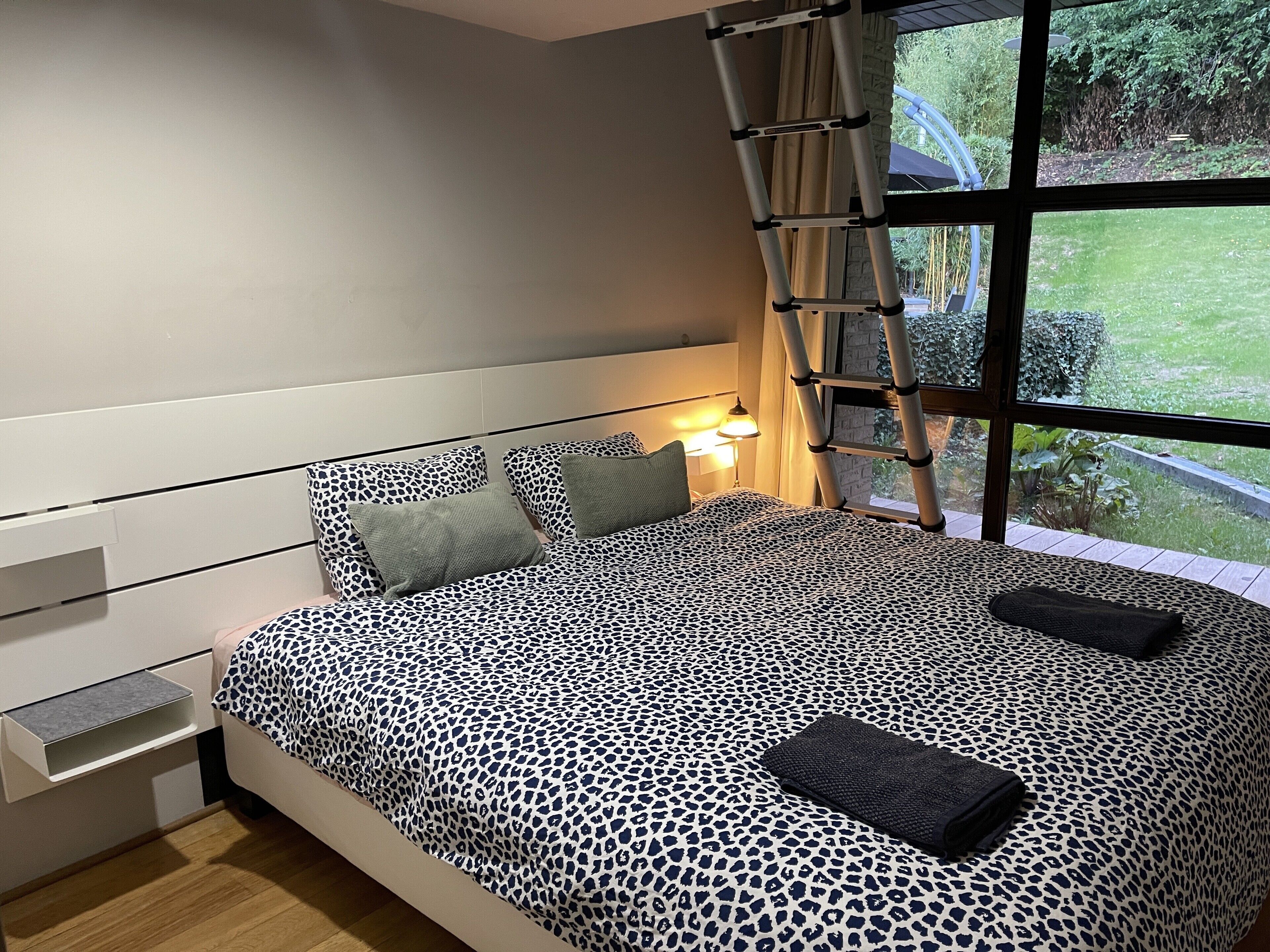 Quarto com cama, travesseiros, mesa de cabeceira, abajur, janela com vista para vegetação e escada.