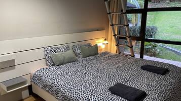 Quarto com cama, travesseiros, mesa de cabeceira, abajur, janela com vista para vegetação e escada.