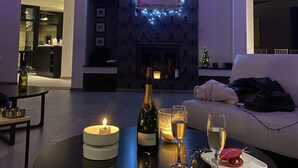 A modern living room with a fireplace, a dining table set for two with a bottle of champagne, a candle, and a dessert.
