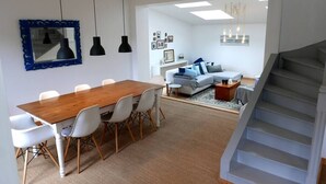 A modern living room with a wooden dining table, white chairs, a grey sofa, and a staircase. - Village house Scandinavian charm (Carantec)