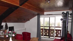 Un salon chaleureux avec une table en bois, des chaises rouges, un banc en bois et un canapé rouge. Il y a une fenêtre donnant sur l’extérieur et un ventilateur de plafond.