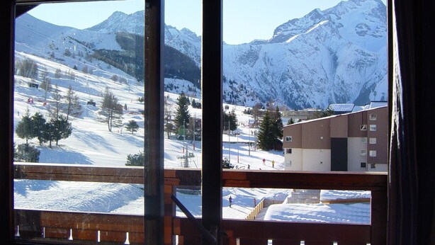 Une vue sur une chaîne de montagnes enneigées, aperçue à travers une fenêtre, avec une station de ski et des bâtiments visibles au premier plan.