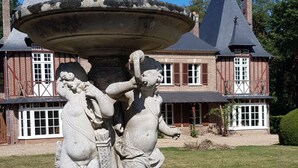 A stone fountain with two figures, a brick building with a gabled roof, and a well-maintained lawn. - Manoir du Chambellan (Les Baux-Sainte-Croix)