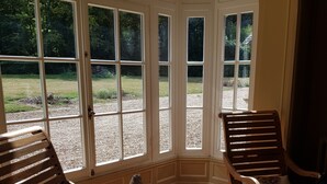 A sunlit room with wooden furniture, including two chairs and a small table, and large windows offering a view of a garden. - Manoir du Chambellan (Les Baux-Sainte-Croix)