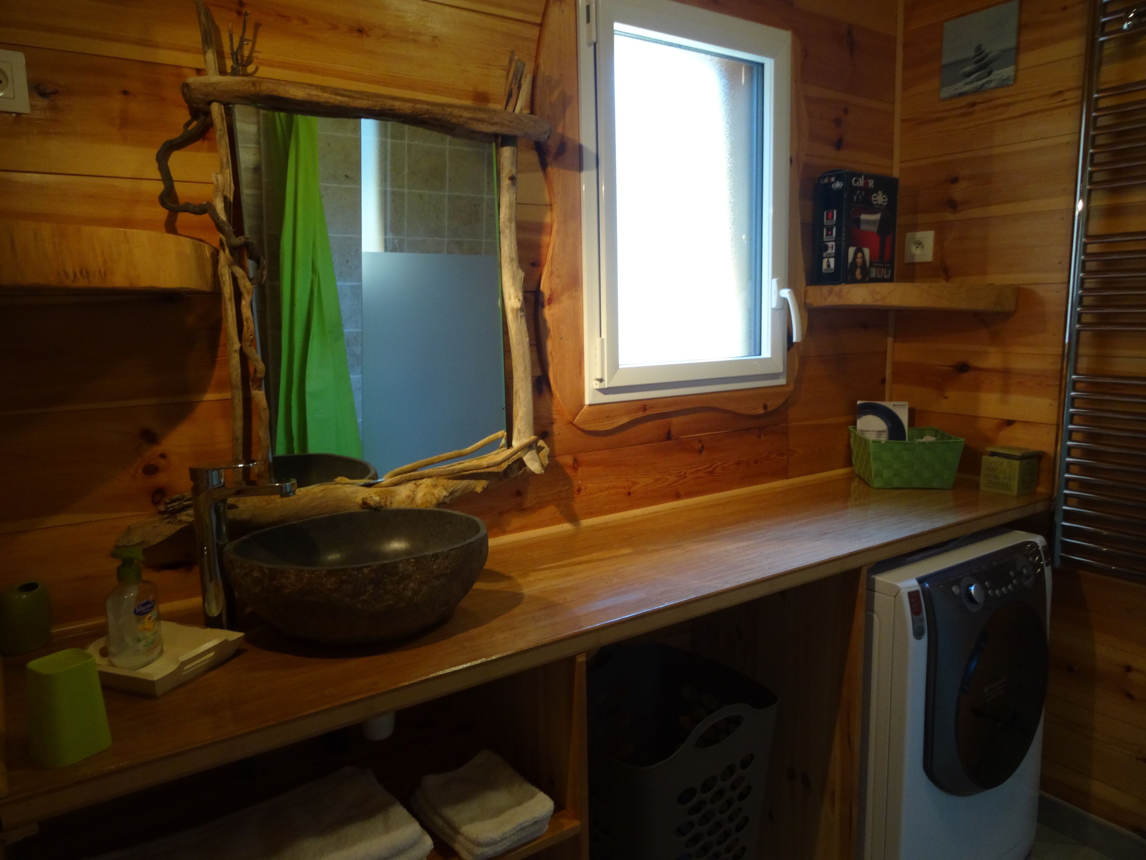 A wooden cabin interior with a stone basin sink, a window with a view, a washing machine, and a green basket on the counter.