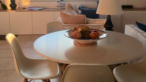 A dining area with a round table, white chairs, and a bowl of fruit. A sailboat model is on the windowsill, and a TV is mounted on the wall. - 2 villas together for an exceptional view with private pool (SKIATHOS)