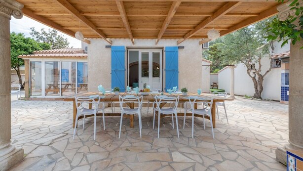 Un patio al aire libre con una pérgola de madera, suelo de piedra y una mesa de comedor con sillas blancas.
