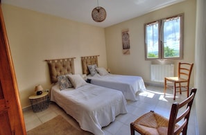 Quarto com duas camas, uma cadeira, mesa de cabeceira, janela com cortinas e um objeto decorativo na parede.