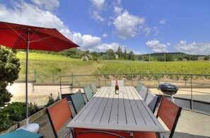 Área de refeições ao ar livre com mesa, cadeiras, um guarda-sol vermelho e vista para um vinhedo.