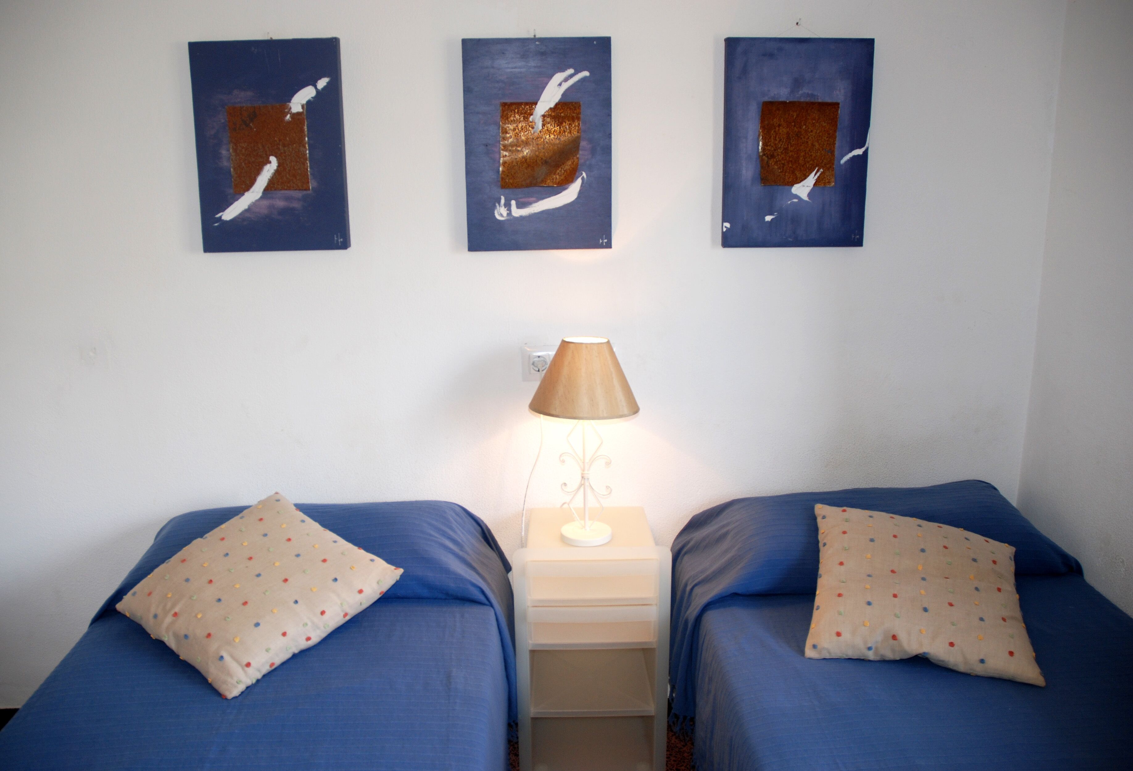 A room with two beds, each with a blue bedspread and a polka-dotted pillow. There is a nightstand with a lamp between the beds. Above the nightstand, there are three framed artworks with abstract designs.