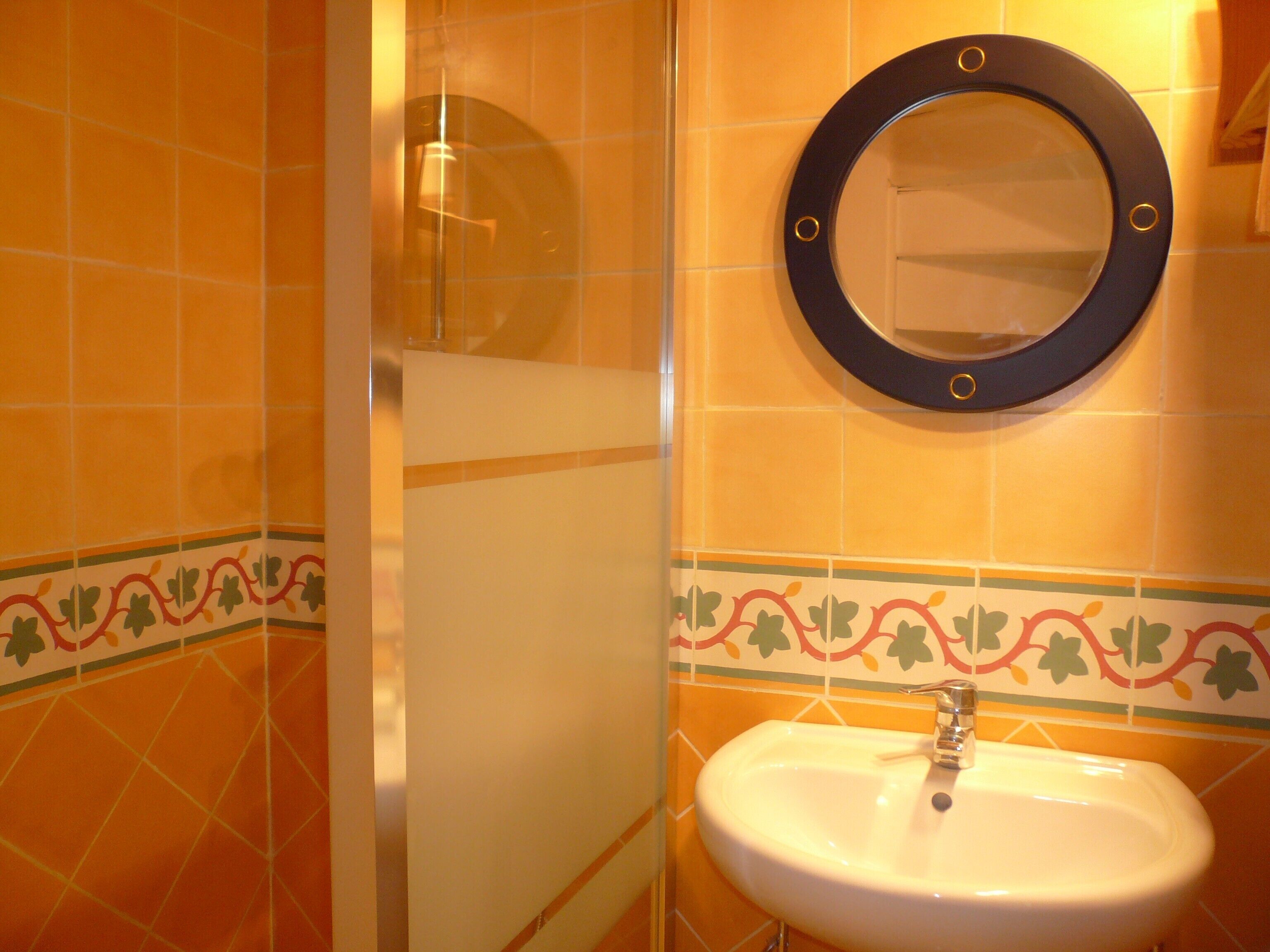 Un baño con paredes de azulejos naranja, un lavamanos blanco y un espejo redondo con marco negro.