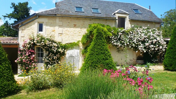 Una casa de pedra amb teulada inclinada, dues petites finestres i un jardí ple de flors i vegetació.