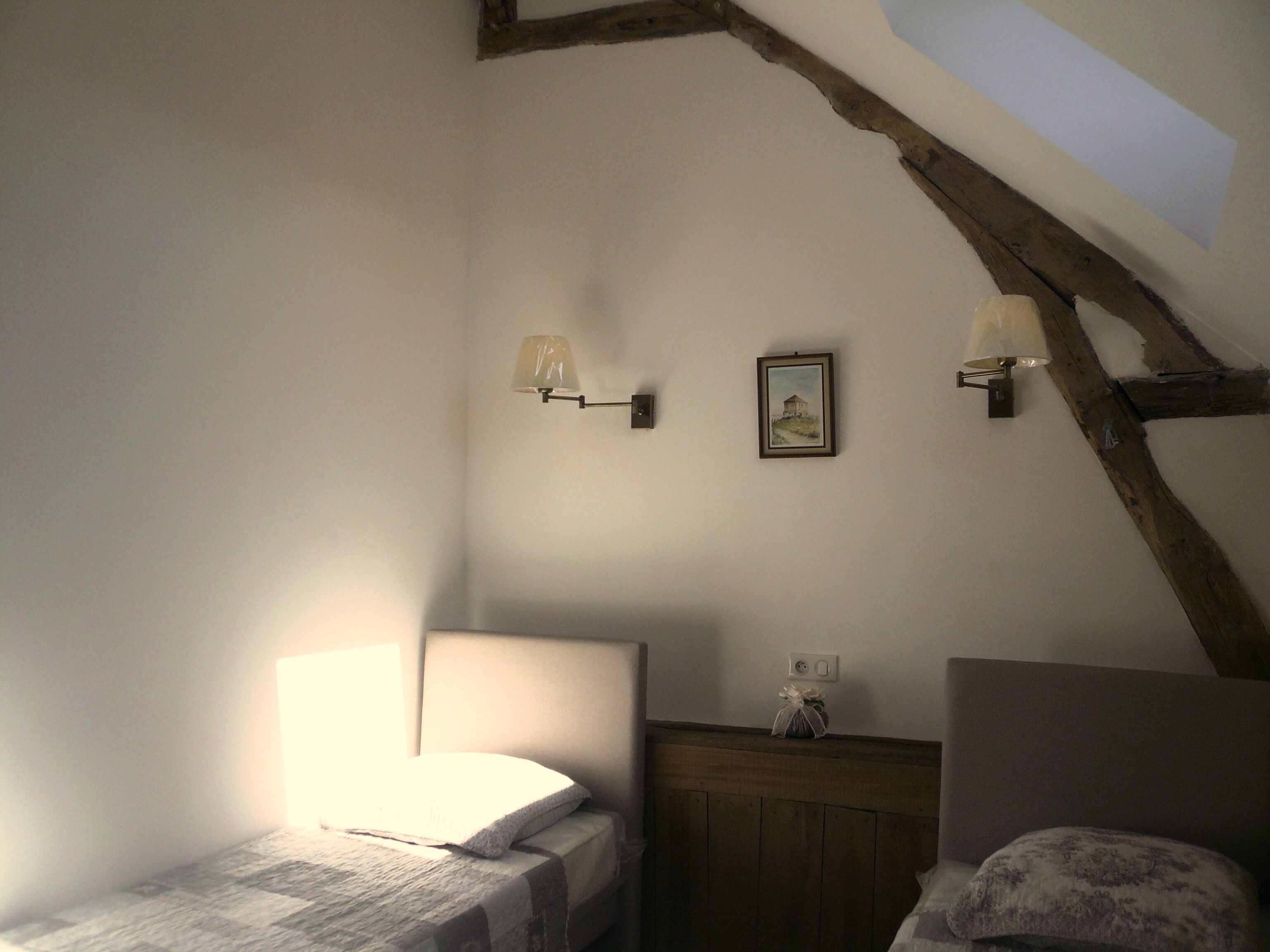 Un dormitorio con techo inclinado, una cama con cabecera blanca, dos lámparas fijadas a la pared y un cuadro enmarcado.