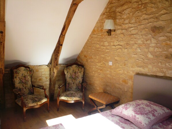 A rustic room with stone walls, wooden beams, two floral-patterned chairs, a small wooden stool, and a bed with a floral bedspread.