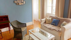 A living room with a blue wall, a white sofa with a blue pillow, a wicker chair, a glass coffee table, and a wooden desk. - Charming apartment in the city center of 63 m2 (Villers-sur-Mer)