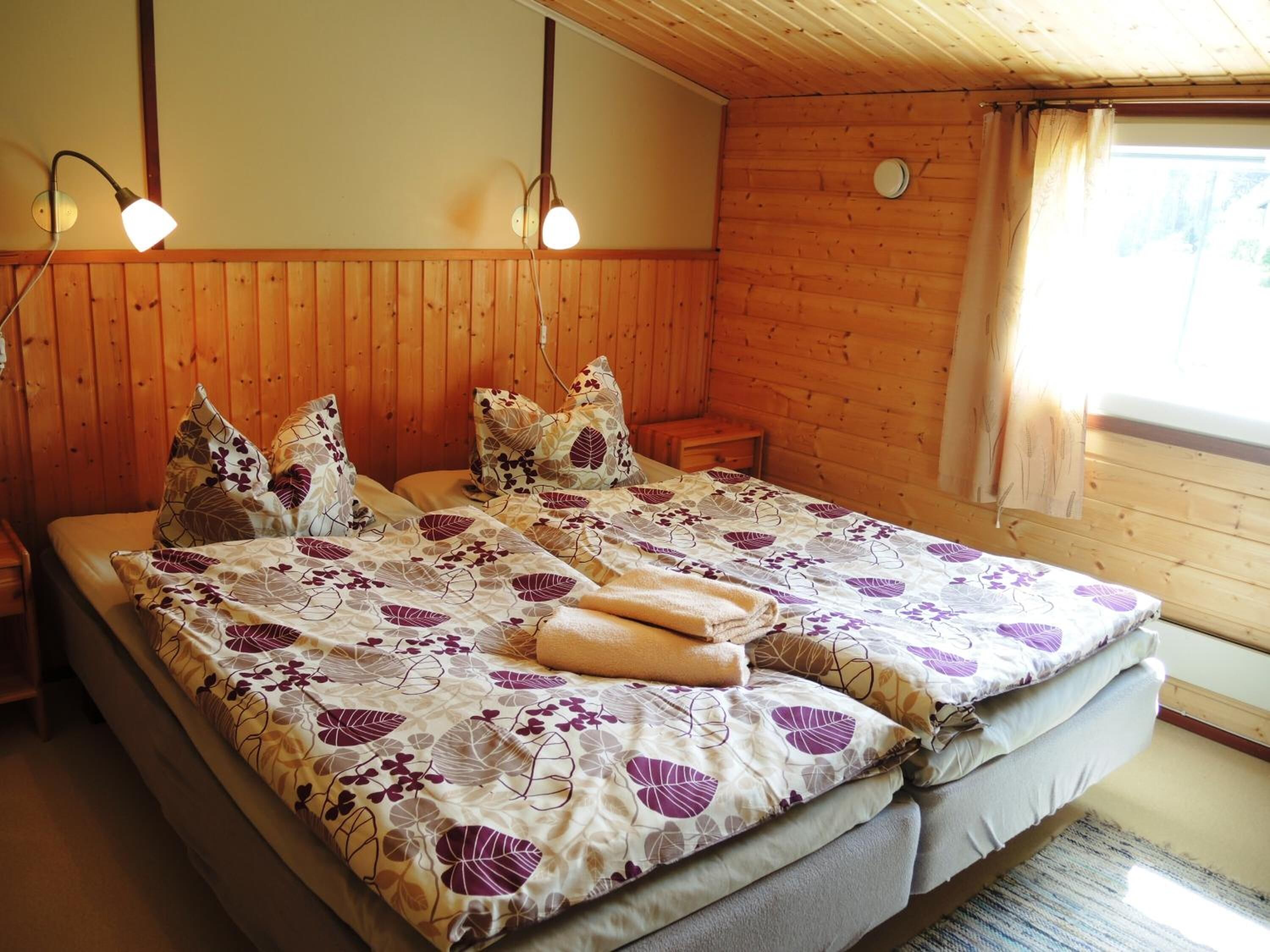 Cama doble con ropa de cama floral, dos lámparas de noche, un cabecero de madera y una ventana con cortinas.