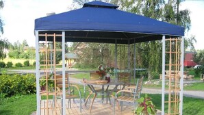 A gazebo with a blue canopy, wooden flooring, and metal railings. It includes a table and chairs.