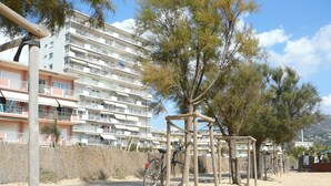 Uma área de praia arenosa com uma estrutura de madeira, possivelmente um parquinho, e um edifício de vários andares com varandas ao fundo.