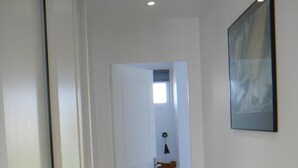 Um corredor estreito com uma porta que dá para um quarto. O quarto possui uma cama, uma mesa de cabeceira e uma luminária fixada na parede.