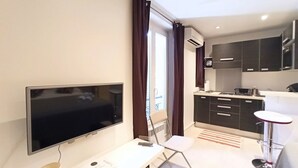 Une chambre d’hôtel moderne équipée d’un téléviseur à écran plat, d’une chaise pliante, d’une petite table, d’une kitchenette et d’une porte-fenêtre coulissante donnant sur le balcon.