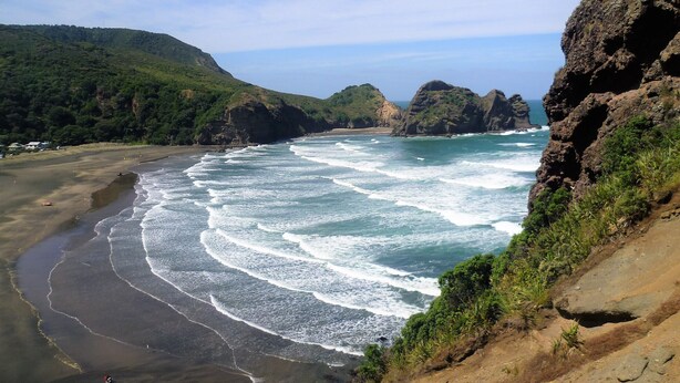 A coastal landscape with a curved sandy beach, rocky cliffs, and lush greenery.