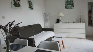 Ein modernes Wohnzimmer mit einer Couch, einem kleinen runden Tisch und einer weißen Kommode. An der Wand hängen zwei gerahmte Bilder, eines zeigt ein Telefon, das andere ein Gebäude.