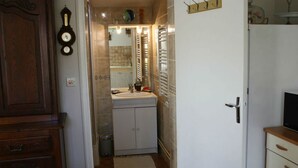 A narrow hallway with a wooden cabinet on the left, a white door on the right, and a bathroom with a sink and mirror in the center. - GITE LACCALMIE 17 - MATHE BRILLOUET CLAUDINE ET PATRICE (Saintes)