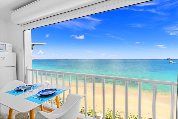 A balcony with a table set for two, overlooking a beach with clear blue water and a boat in the distance.