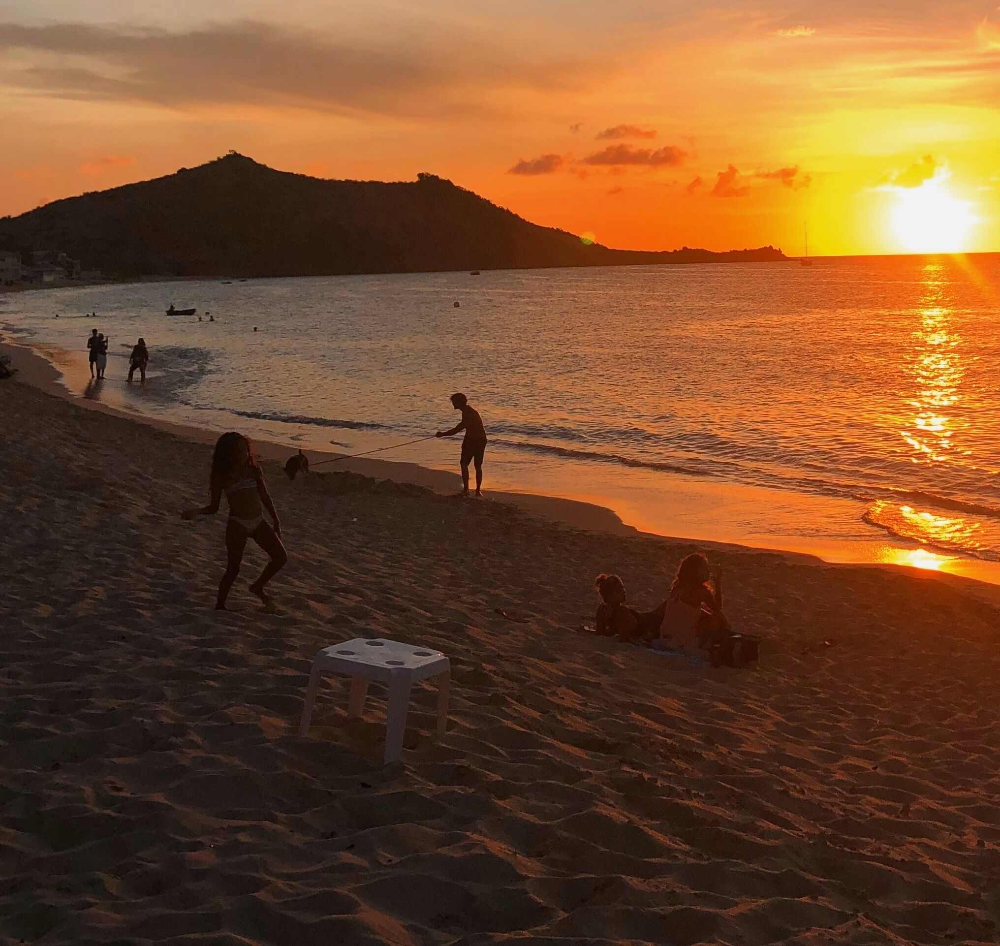 A beach scene with people walking and sitting, a small table, and a sunset over the water.