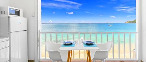 A dining area with a view of the sea, a white table, two white chairs, and a microwave on a white cabinet.