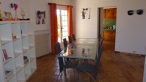 A dining area with a glass table and chairs, a bookshelf, a radiator, and a kitchen visible through an open door.