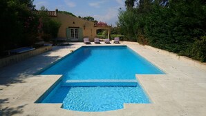 A rectangular swimming pool with a clear blue water, surrounded by a concrete deck. There are lounge chairs aligned along the pool's edge.