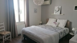 Una camera da letto con un letto, comodini, una sedia, una finestra con tende, un condizionatore d'aria e una mensola a parete con opere d'arte.