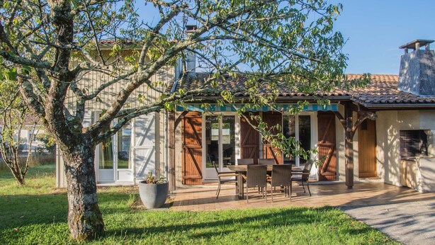 Une maison avec un patio, un coin repas et un grand arbre au premier plan.