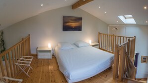 Une chambre avec un plafond en pente, un lit avec une literie blanche, une table de chevet, une chaise en bois, une petite table, une plante et un tableau représentant un coucher de soleil.