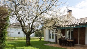 Une maison avec un jardin, une terrasse avec une table et des chaises, et un arbre aux fleurs blanches.