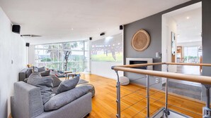 A modern living room with a grey sofa, wooden flooring, a TV displaying a sports event, and a balcony with a view. - Villa, heated pool, exceptional view, exotic garden, 4-star rating (Montbazin)