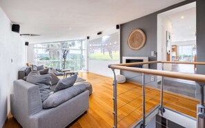 A modern living room with a grey sofa, wooden flooring, a TV displaying a sports event, and a balcony with a view. - Villa, heated pool, exceptional view, exotic garden, 4-star rating (Montbazin)
