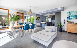 A modern living room with a sofa, dining area with chairs, a kitchen, and a large window. - Villa, heated pool, exceptional view, exotic garden, 4-star rating (Montbazin)
