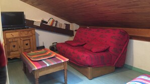 A cozy attic room with a wooden ceiling, a red patterned sofa, a small wooden table, a television, and a bookshelf. - Apartment in the center of the village of Combloux (Combloux)