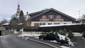 A two-story wooden building with a balcony, a stone wall, and snow on the ground. - Apartment in the center of the village of Combloux (Combloux)