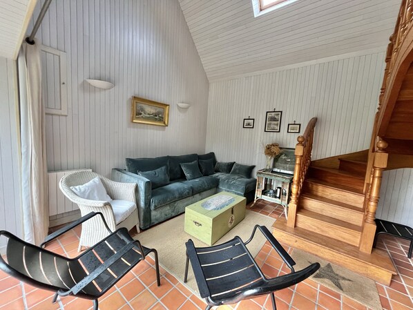 A cozy living room with a green sofa, white armchairs, and a coffee table. A staircase with wooden railings leads to an upper level.