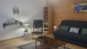A living room with a dark blue sofa, a wooden bookshelf, a chair with a palm tree cushion, a small table, and a wall-mounted shelf with decorative items. - Les balcons de Fellering: Spacious apartment with panoramic view (Fellering)