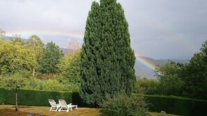 A garden with a tall coniferous tree, two white lounge chairs, and a rainbow in the background. - Les balcons de Fellering: Spacious apartment with panoramic view (Fellering)