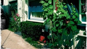 Una casa de fachada verde, tejado marrón y chimenea. Cuenta con un jardín con plantas verdes y flores rojas.