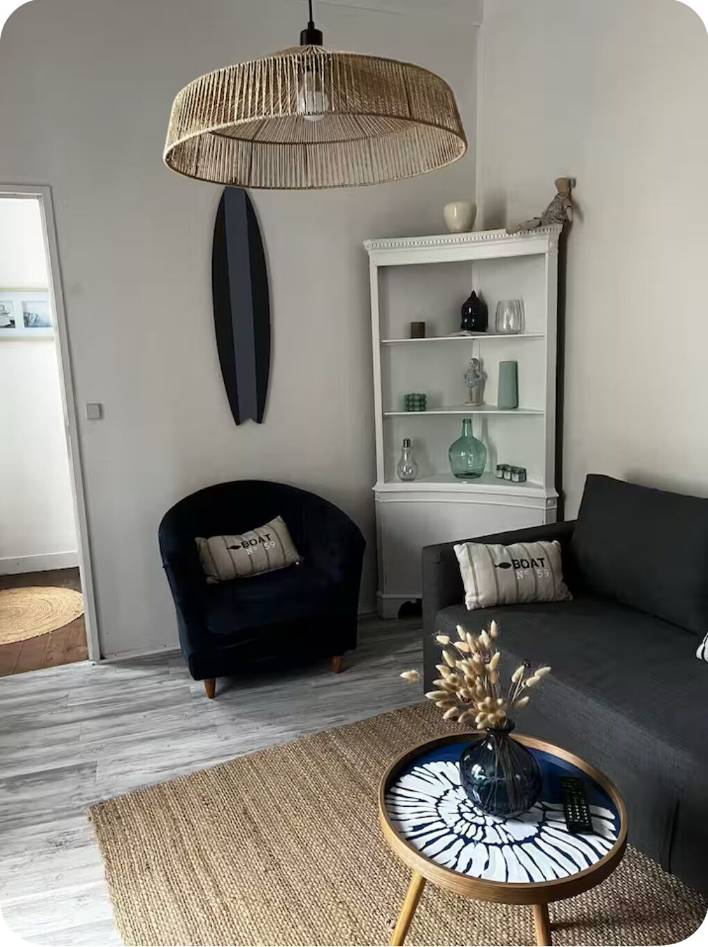 En moderne stue med en mørkeblå lænestol, en grå sofa, et hvidt reolsystem med dekorative genstande, en vævet gulvlampe og et rundt sofabord med en vase.
