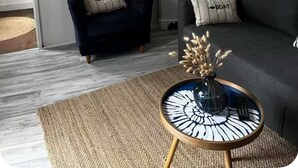 Een moderne woonkamer met een donkerblauwe fauteuil, een grijze bank, een wit wandrek met decoratieve objecten, een geweven vloerlamp en een rond bijzettafeltje met een vaas.