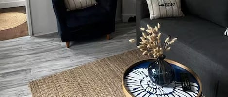 En moderne stue med en mørkeblå lænestol, en grå sofa, et hvidt reolsystem med dekorative genstande, en vævet gulvlampe og et rundt sofabord med en vase.