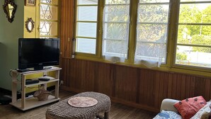A living room with a television on a stand, a wicker ottoman, a patterned sofa, and a window with lace curtains.