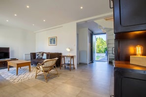 A modern living room with a sofa, a coffee table, a TV, and a wicker chair. There is a painting on the wall and a door leading outside.